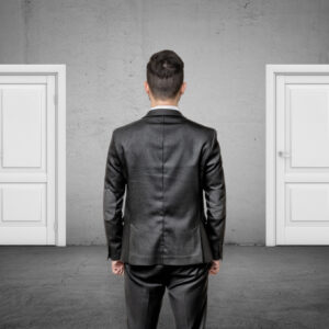 A businessman with his back turned stands between two identical closed white doors. Make right choice. Important decision. Never fail.
